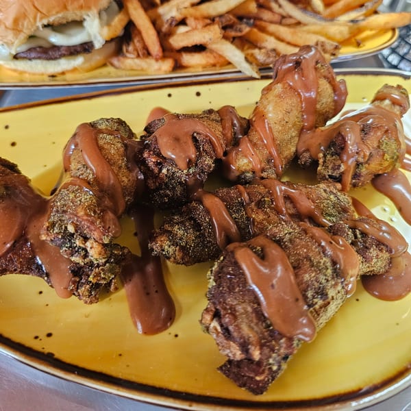 Chocolate-drizzled churro-style fried chicken wings on a yellow plate, with a burger and fries in the background.