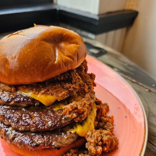 A towering Urban Burger with multiple patties, melted cheese, and chili, on a pink plate.