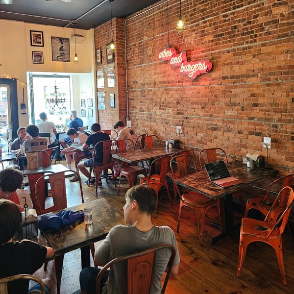 Diners seated at rustic tables with orange chairs in a burger restaurant with brick walls and neon sign.