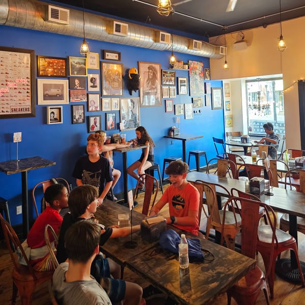 Diners, including several children, seated at rustic tables in Urban Burger, with a vibrant blue wall adorned with eclectic art.