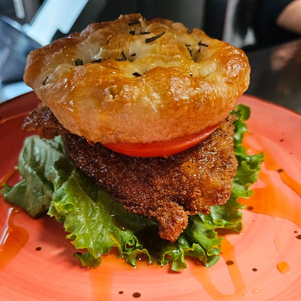A crispy fried chicken sandwich on a focaccia bun with lettuce and tomato, drizzled with sauce.