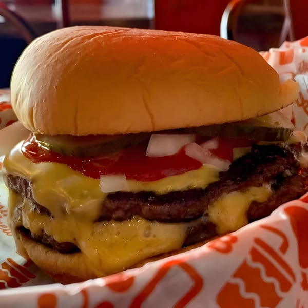 Close-up of a double cheeseburger with melted cheese, ketchup, onions, and pickles in a paper wrapper.