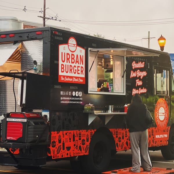 A person stands in front of the black Urban Burger food truck with its service window open.
